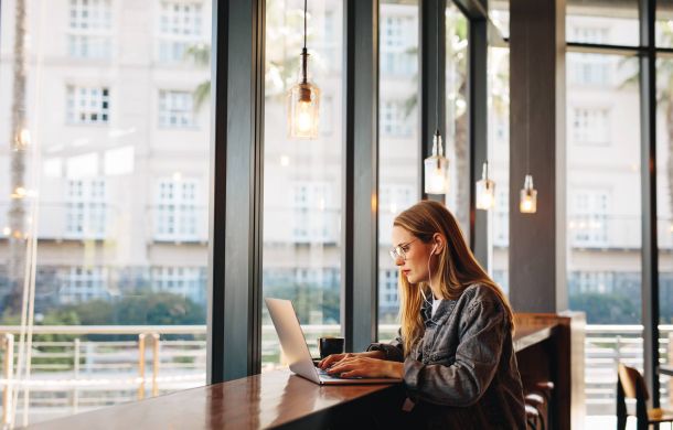 Woman in glasses working on a laptop at a window-side counter in a bright, modern café.