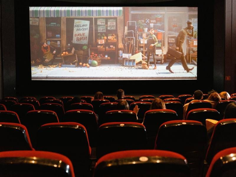 People watch an animated movie in a theater with red seats and a scene of a street shown on the screen.
