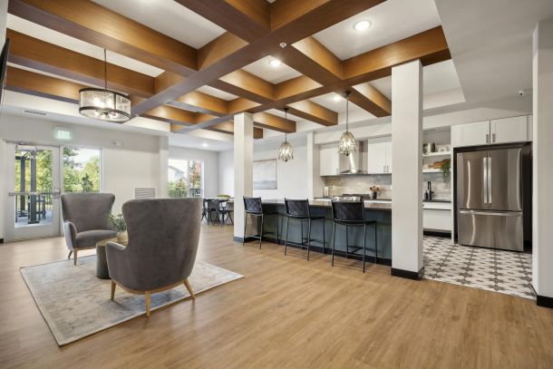 Modern open kitchen and lounge area with wood floors, pendant lights, and gray chairs near large windows.