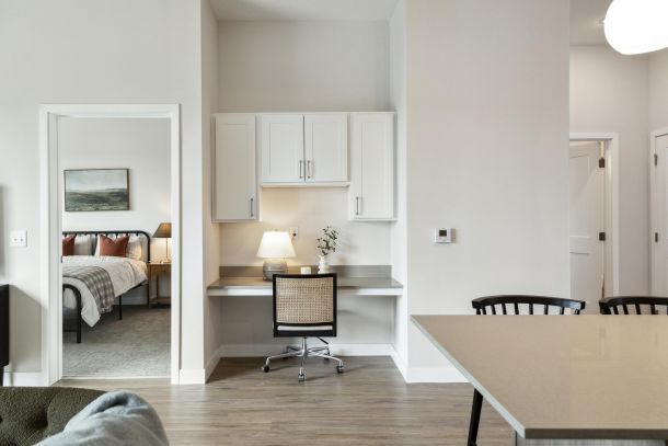 A home office nook with a desk, chair, lamp, and cabinets, next to a bedroom and modern dining area.