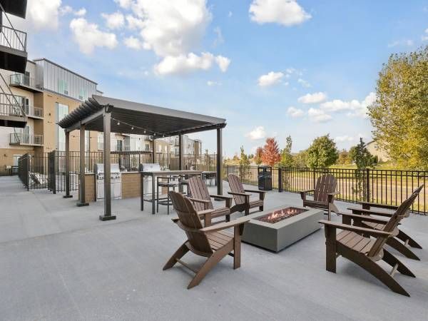 Outdoor patio with wooden chairs around a fire pit, pergola, and grill area, next to a building and trees.