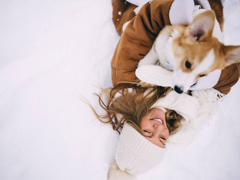 Woman in winter clothes lying in the snow, smiling, and hugging a corgi dog.
