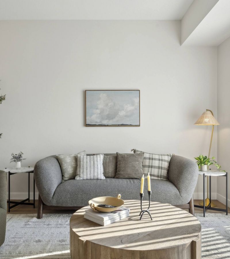 Modern living room with a gray sofa, round wooden table, and natural light from large window with vertical blinds.