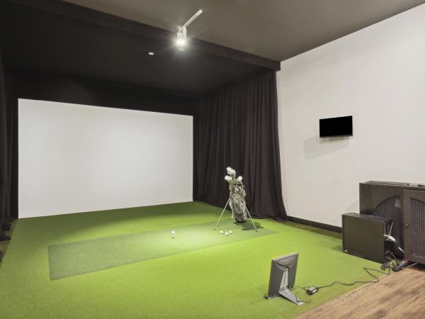 Indoor golf simulator room with a screen, golf clubs, turf mat, and various equipment.