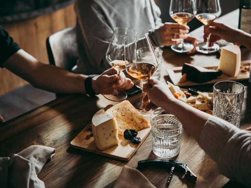 Four people clink wine glasses over a table with assorted cheeses and snacks on wooden boards.