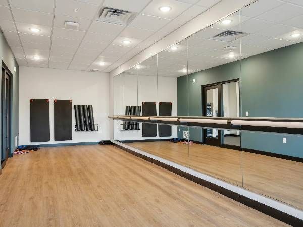 Empty exercise studio with wood floor, mirrored wall, ballet barre, and stacked mats in the corner.