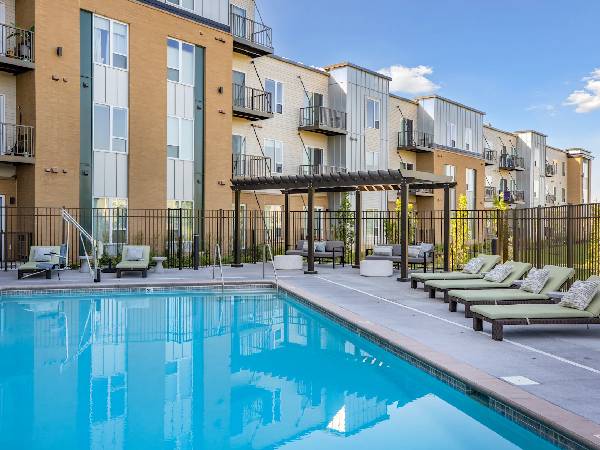 Modern apartment complex with pool, lounge chairs, and shaded seating area under a clear blue sky.
