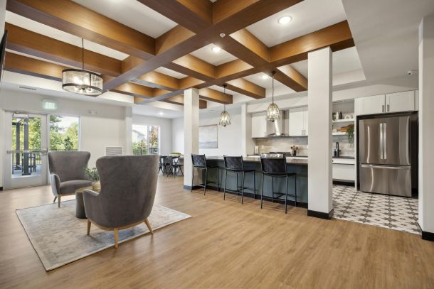 Modern open kitchen and lounge area with wood floors, pendant lights, and gray chairs near large windows.