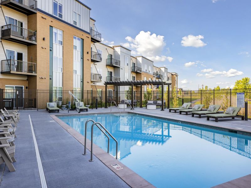 Modern apartment complex with outdoor swimming pool, lounge chairs, and partly cloudy sky in the background.