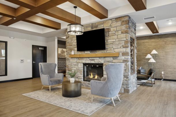 Modern lounge with stone fireplace, two gray chairs, round table, and wood-beam ceiling.